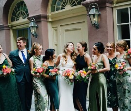 Bride laughs with bridesmaids and groomsman in front of arched windows, holding colorful bouquets at a New Orleans wedding.