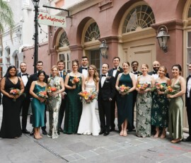 Wedding party of 18 smiles outside a historic Louisiana building; bridesmaids in green, groomsmen in black suits, holding bouquets.