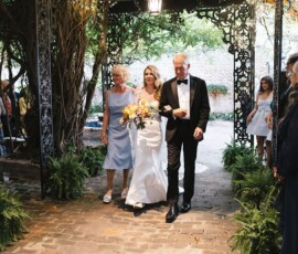 Bride in white dress walks with two older adults down an elegant outdoor aisle at a classic New Orleans wedding.