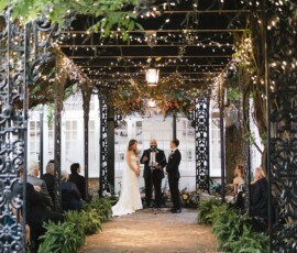 A couple weds under a vine-covered gazebo with string lights, surrounded by guests at a romantic Gulf South outdoor ceremony.