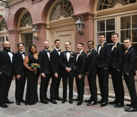 Wedding party in black formal wear poses and smiles outdoors by a historic building, capturing Gulf South elegance.
