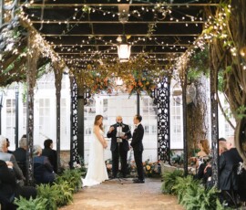 Couple exchanges vows under pergola with string lights, surrounded by guests, greenery, and ferns on a brick pathway.