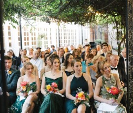 Wedding guests and bridesmaids in green dresses with bouquets sit outdoors under leafy vines and string lights in Louisiana.
