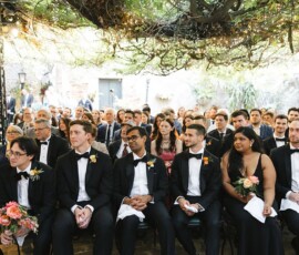 Guests in tuxedos and formal dresses sit under a vine-covered canopy at an elegant New Orleans wedding.