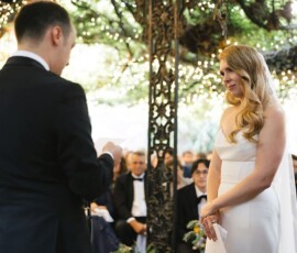 A bride smiles as the groom reads vows at an outdoor wedding, guests seated under string lights in the background.