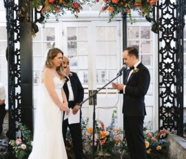 Bride and groom under floral arch as groom reads vows; smiling bride, officiant, and elegant flowers evoke Gulf South wedding charm.