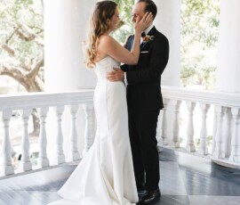 Bride and groom smile and touch faces on a porch with white columns and trees, creating elegant wedding inspiration.