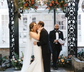 Bride and groom kiss under a floral arch at a classic New Orleans wedding, with the smiling officiant standing behind them.
