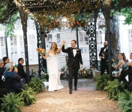 Smiling bride and groom walk hand in hand under a decorated gazebo at a Louisiana wedding, surrounded by greenery and flowers.