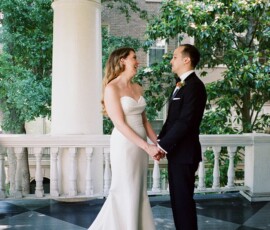 Bride and groom hold hands on a porch, smiling amid greenery and white columns at a classic New Orleans wedding.