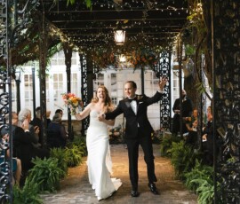 Bride and groom walk hand in hand down a vine-covered outdoor aisle, smiling, with guests seated on both sides.