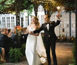 Smiling bride and groom walk hand-in-hand down an outdoor aisle under string lights at their Louisiana wedding.