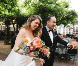 Bride with vibrant bouquet walks hand-in-hand with groom at outdoor Louisiana wedding as guests look on.
