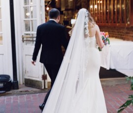 Bride in white gown and veil walks with man in black suit to a New Orleans wedding venue, bouquet in hand, before ceremony.
