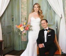 Bride in white gown with bouquet stands by groom in tux on orange chair, both smiling in a vintage Louisiana wedding room.