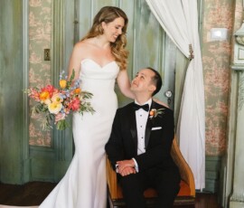Bride in white gown with bouquet stands by seated groom in tux; both smile in vintage room with green accents and floral wallpaper.