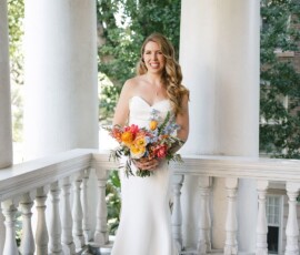 Smiling bride in a white strapless gown holds a colorful bouquet on a porch with white columns and lush New Orleans greenery.