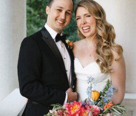 Smiling wedding couple on a Louisiana porch with bouquet, green trees, and white columns in the background.