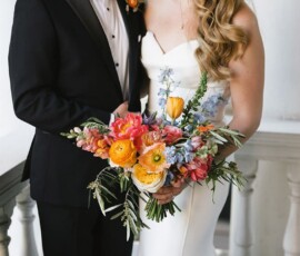 Bride in white dress with bouquet stands by person in black suit on porch, perfect Gulf South wedding inspiration.