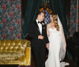 Bride in white dress and veil holding groom’s hand, smiling at Louisiana wedding by gold sofa and green curtains.