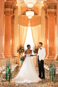 Bride in white gown and groom in white jacket stand together in an ornate hall with gold chairs, chandeliers, and flowers.