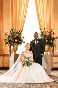 Bride in white gown with bouquet sits as groom in black tuxedo stands beside her, posing by gold vases and curtains.