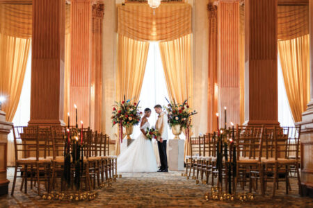 Bride and groom at altar in elegant gold hall with tall pillars, chairs, floral arrangements, and candles along the aisle.