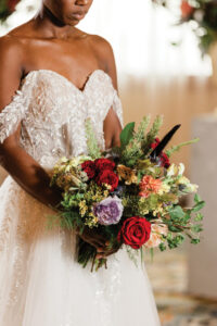 Bride in off-shoulder white lace gown holding a vibrant bouquet of red, purple, yellow, peach flowers, and greenery.