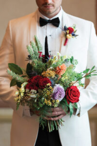 Man in a white tuxedo jacket and black bow tie holds a bouquet with red roses, purple flowers, yellow accents, and greenery.