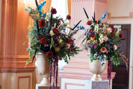 Gold vases with red roses, blue flowers, and greenery on pedestals in an elegant peach-walled room.