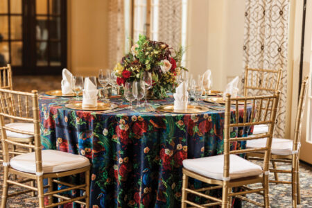 Elegant round table with dark blue floral tablecloth, gold chairs, wine glasses, and a large flower centerpiece.