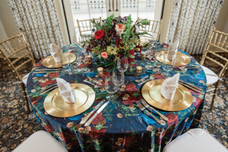 Elegant round table with dark floral cloth, gold chargers, white napkins, colorful centerpiece, and gold chairs.