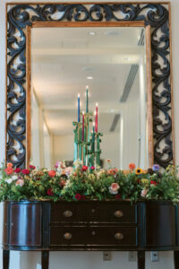 Ornate mirror over dark wood dresser with colorful flowers and three lit candles of various colors in the center.