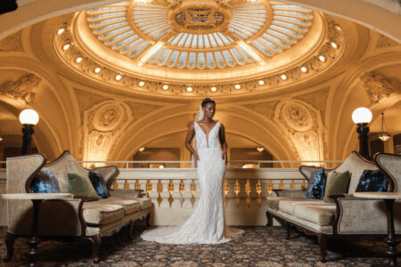 Bride in white gown stands between sofas beneath a grand domed ceiling with lights in a luxurious setting.