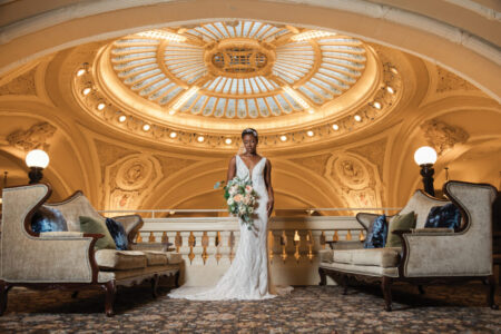 Bride in a white gown with bouquet stands between sofas beneath a grand, ornate dome ceiling with warm lighting.