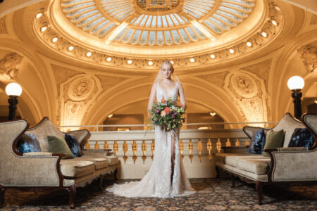 Bride in lace gown with bouquet stands between ornate sofas beneath grand domed ceiling in elegant historic venue.