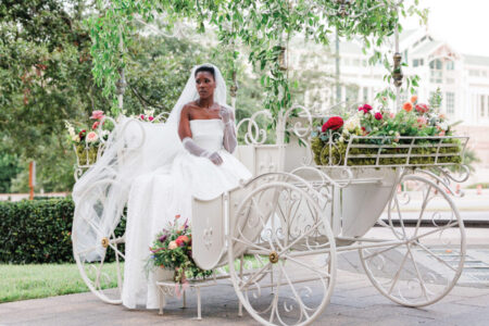 Bride in white dress and veil sits in ornate flower-adorned carriage, with greenery and buildings behind her.