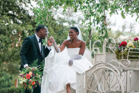 Bride in white carriage and groom in suit smiling and holding hands, with bouquet and greenery decorating the carriage.