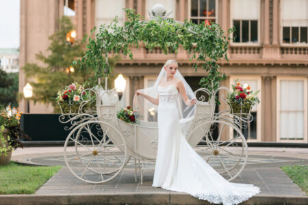Bride in white gown and veil stands by a floral-decorated carriage outdoors near elegant buildings.