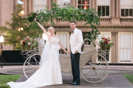 Bride and groom in wedding attire pose before a decorative carriage outdoors; bride smiles, holding bouquet up.