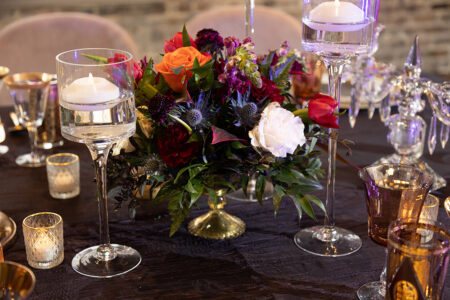 Floral centerpiece with red, orange, purple, and white blooms on a dark wood table with candles and gold votive holders.