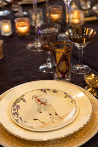 Elegant dinner table with gold-edged plates, glasses, candles, and a top plate showing a camel rider and purple flowers.