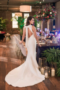 Woman in white backless gown with bow stands by elegant table with flowers, candles, and glassware in a stylish, bright room.