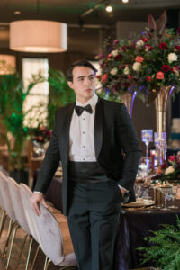 Man in black tuxedo by a formally set dining table with flowers, candles, and tableware in an elegant setting.