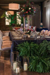 Elegant dining table with formal settings, tall floral centerpieces, glass candle holders, and green ferns on the floor.