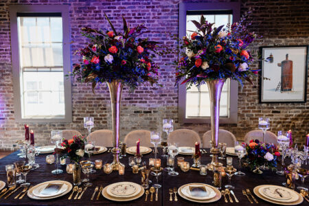 Elegant dining table with plates, cutlery, candles, and vibrant flowers in silver vases, set by brick walls and large windows.