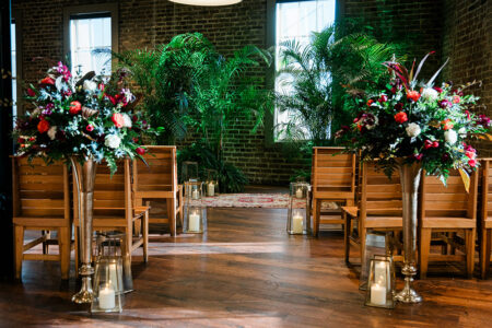 Wooden chairs face a small altar with flowers, plants, lanterns, and candles in a softly lit room with brick walls and windows.