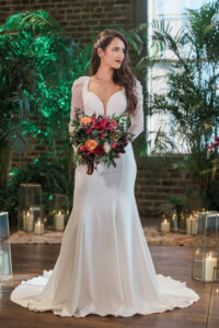 Bride in white gown holding vibrant bouquet indoors with candles, greenery, and brick wall in background.