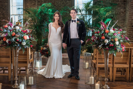 Bride in white gown and groom in black tuxedo hold hands in a flower-filled, candle-lit indoor venue with wooden chairs.