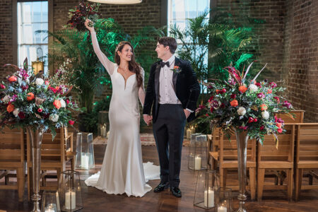 Bride in white dress raises bouquet beside groom; they smile, holding hands in a decorated indoor flower-lined aisle.
